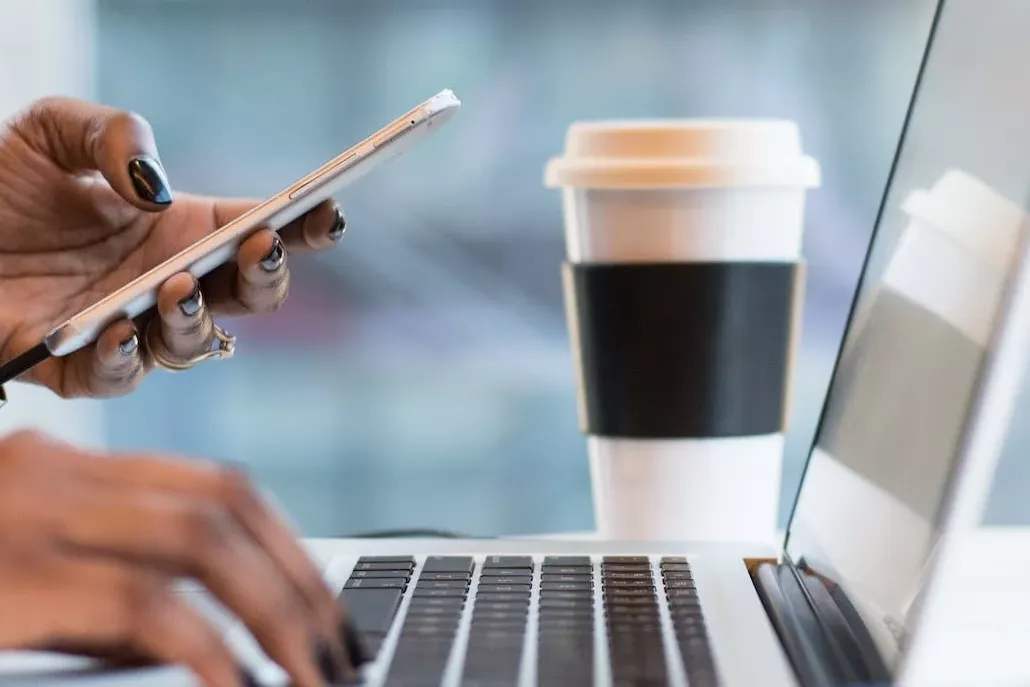 laptop vrouw tikkend met tel in handen met kop koffie lang de kant