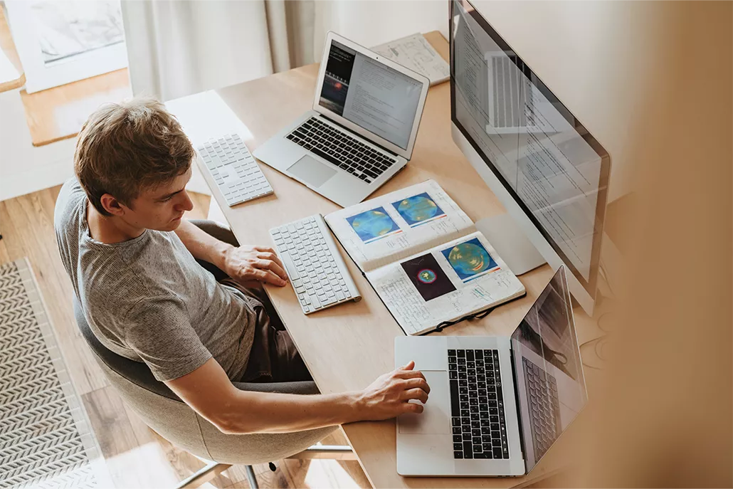 schuin bovenaanzicht, man in shirt werkend aan bureau met een grote Mac en 2 laptops aan weerszijde
